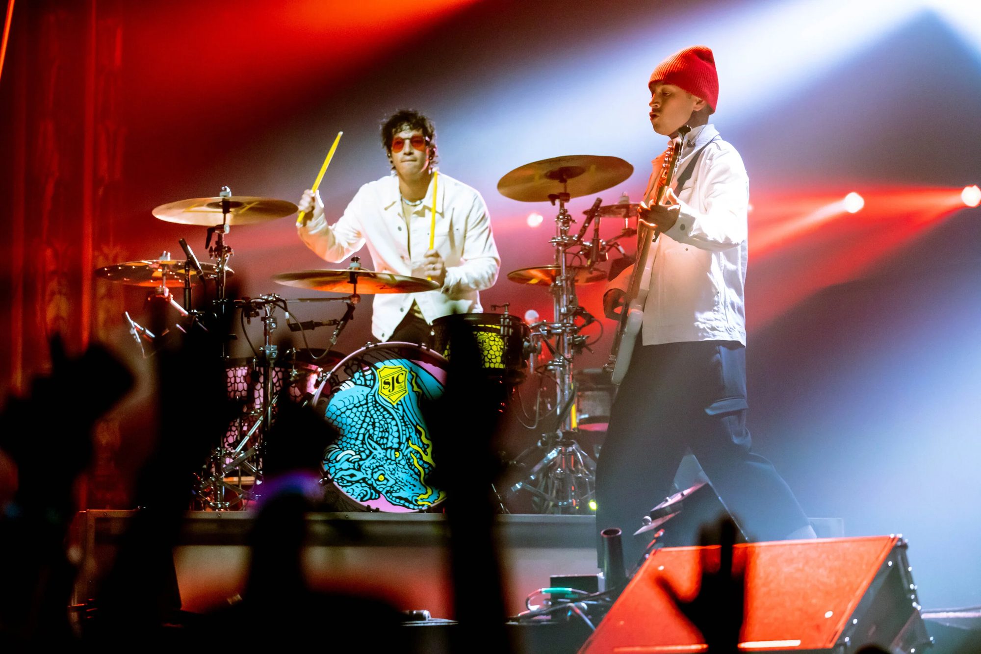 Two performers on stage, one on drums and one with a guitar, under red and blue lights